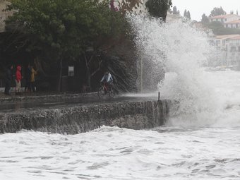 Una onada petant contra el front de mar de Cadaqués, amb vianants mirant el temporal i un ciclista esquivant l'aigua. ANDREU PUIG