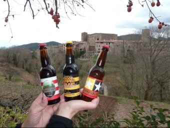 Els tres tipus de cervesa elaborada amb ingredients de Santa Pau, amb el nucli romànic de teló de fons. J.C